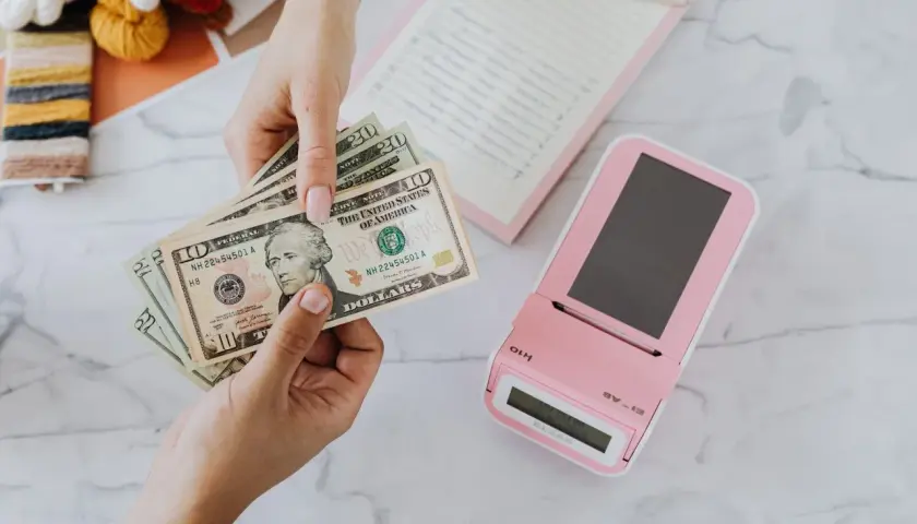 Mani che contano banconote dollari vicino a un registratore di cassa rosa. Un professionista, uno psicologo economico, analizza le spese.