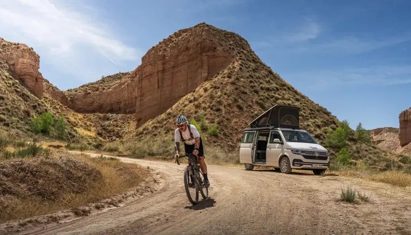 Ciclista pedaleando por un camino de badlands gravel, con una furgoneta camper aparcada al fondo.