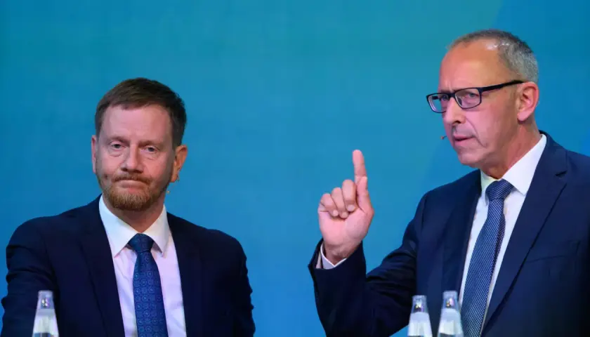 Zwei Männer im Anzug, einer mit Bart, der andere mit Brille und erhobenem Zeigefinger, diskutieren die Wahlprognose Sachsen 2026.