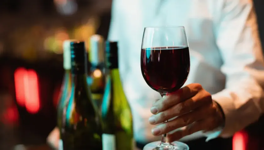 Ein Weinglas mit Rotwein wird gehalten. Im Hintergrund stehen Flaschen. Wo steht das Wasserglas und wo das Weinglas? Hier ist nur das Weinglas zu sehen.