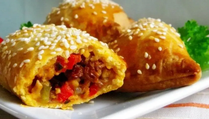 Złociste empanadas posypane sezamem, z widocznym farszem z warzyw i mięsa. Pyszna przekąska.