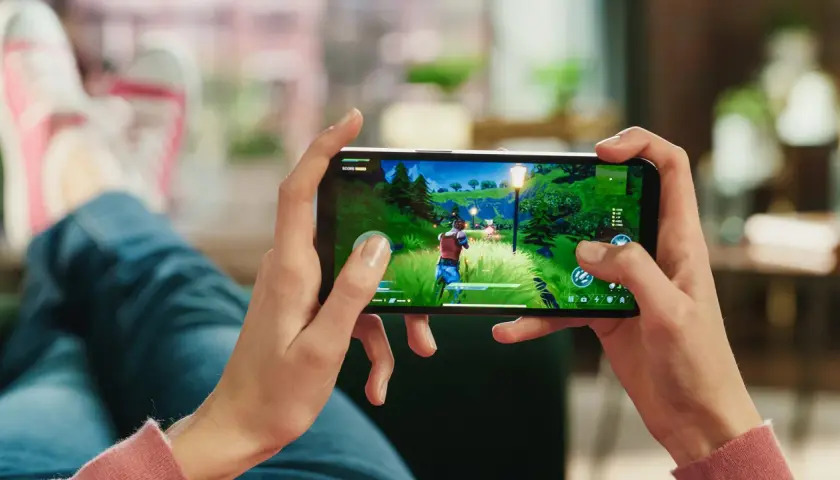 Ręce trzymają smartfon, na którym widać fragment popularnej gry na telefon. W tle widać nogi w jeansach i trampkach.