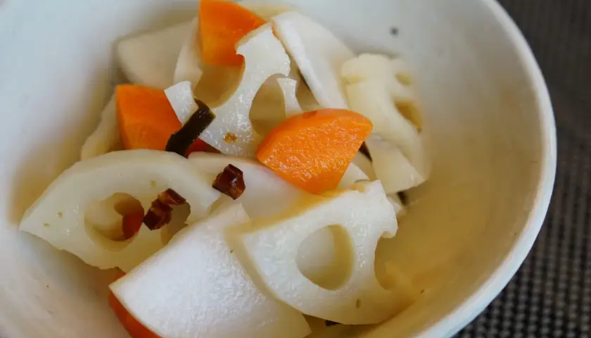 A close-up of a bowl filled with a colorful mix of Japanese root vegetables, including sliced lotus root with its distinctive holes, bright orange carrot pieces, and possibly daikon radish.