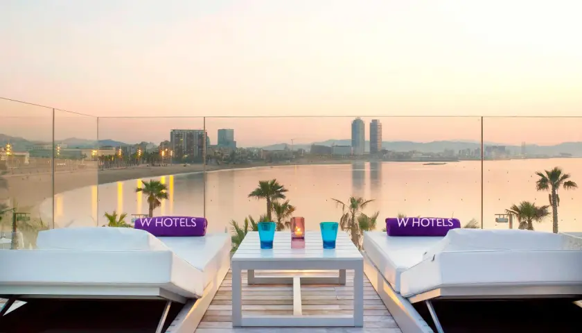 Dos tumbonas blancas con toallas moradas de W Hotels, una mesa con vasos y velas, y vistas al mar y la ciudad de Barcelona al atardecer.