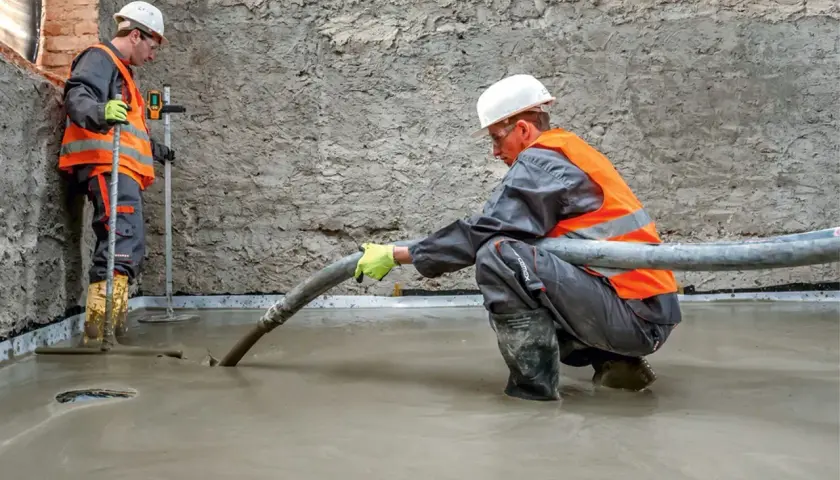 Dwóch budowlańców w kamizelkach odblaskowych wylewa beton. Jeden z nich kieruje wąż, z którego leje się masa, drugi kontroluje poziom. To praca z keramzytem budowlanym.