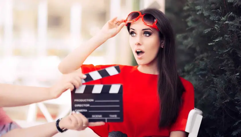 Aktorka w czerwonej sukience z okularami na głowie, zszokowana, gdy klaps filmowy zbliża się do jej twarzy. Stopniowanie high.