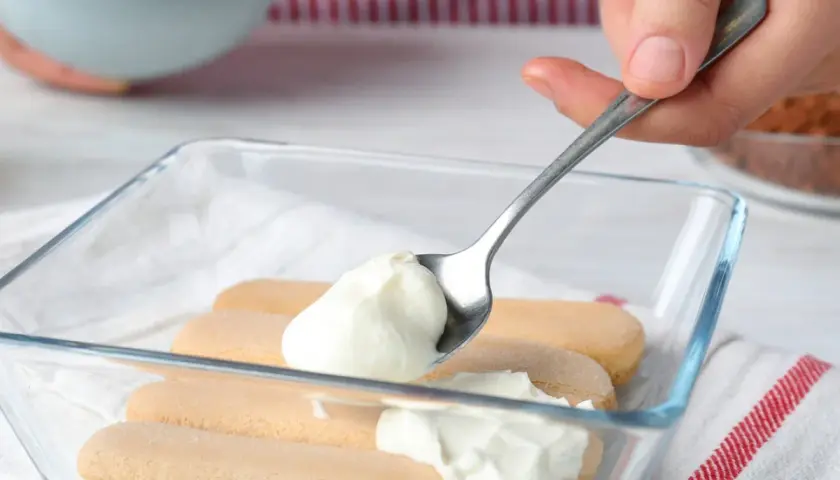 Nakładanie łyżką puszystego kremu z mleka skondensowanego i mascarpone na biszkopty w szklanej formie.