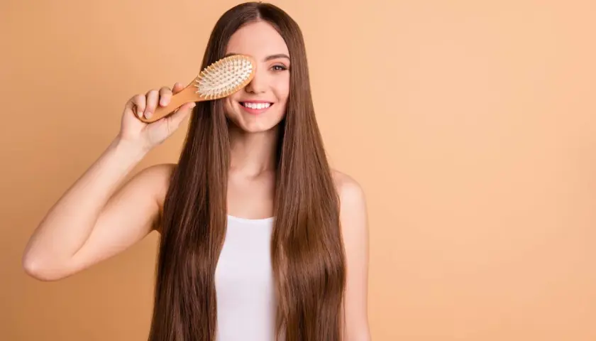 Mujer sonriente con pelo largo y liso, sosteniendo un cepillo de madera sobre su ojo. ¡Un truco para saber como desenredar un pelo muy enredado!