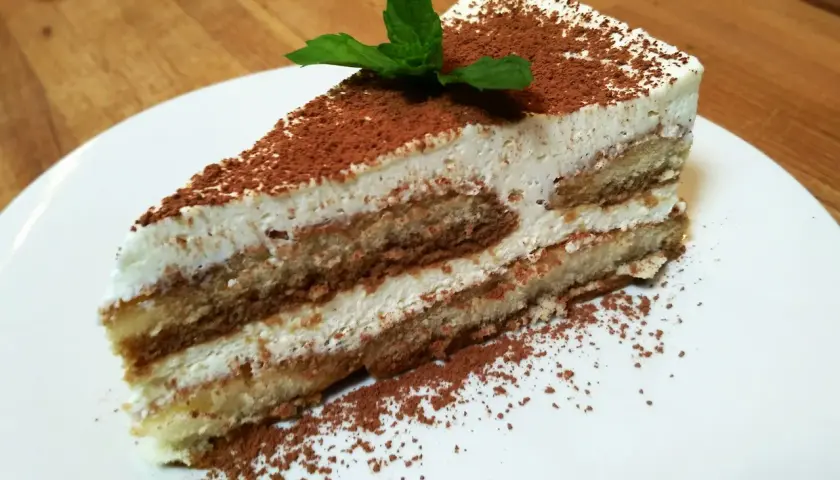 Pyszny kawałek najprostszego tiramisu, posypany kakao i udekorowany listkiem mięty, na białym talerzu.