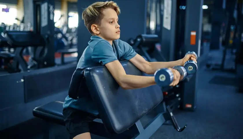 Chłopiec ćwiczy biceps z hantlami na ławce. To jego siłownia dla 12 latka, gdzie buduje siłę.