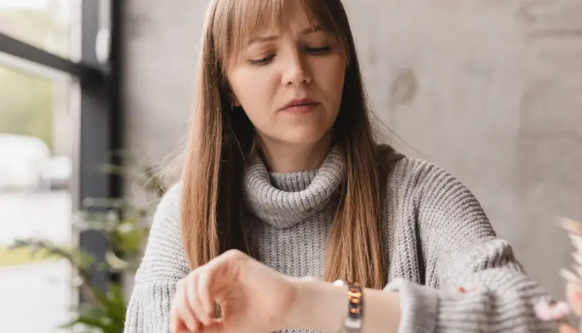 Zamyślona kobieta w szarym swetrze zerka na zegarek. Czyżby randka z aplikacją? 20 20 znaczenie dla singli.