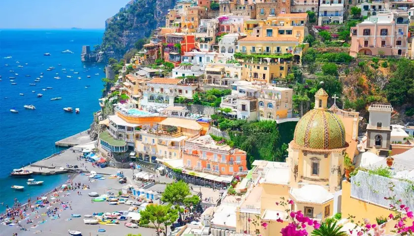 Colorful towns near Naples, Italy, cascade down a cliff to a busy beach and blue sea. A domed church stands out.
