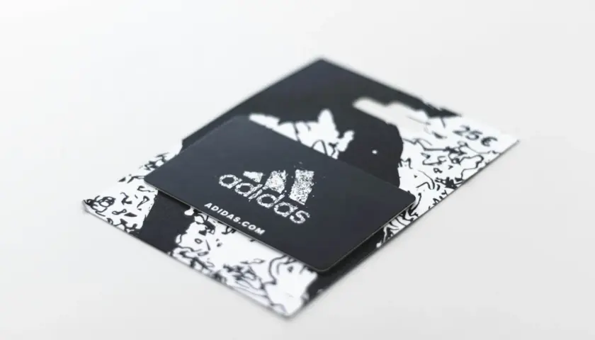 Czarny voucher Adidas z logo i adresem strony internetowej. Dowiedz się, jak zrobić voucher na prezent.