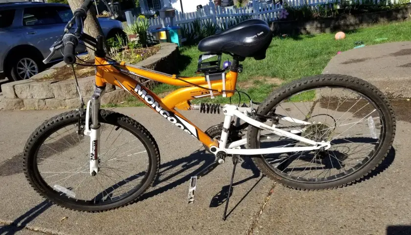 An orange Mongoose XR-75 mountain bike with a black seat and white suspension sits on a sidewalk.