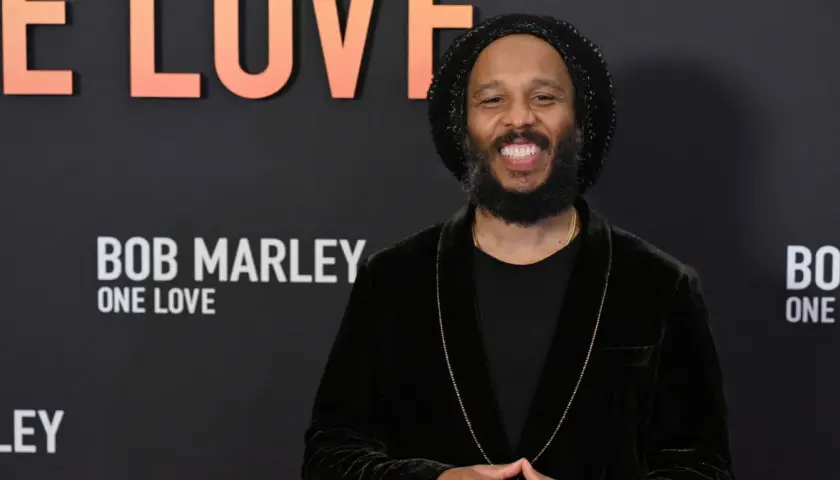 Uśmiechnięty mężczyzna w czapce i aksamitnej marynarce, przypominający Boba Marleya, z napisem "Bob Marley One Love" w tle. Może to być jeden z jego dzieci.