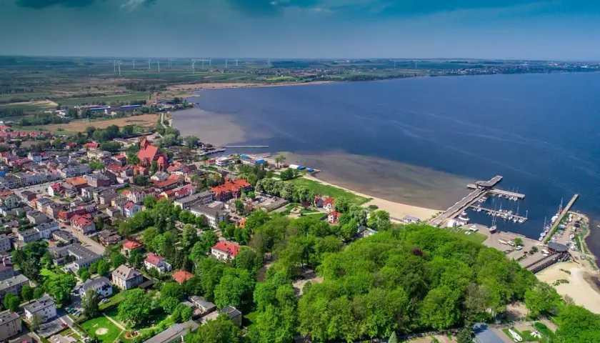 Widok z lotu ptaka na miasto nad Zatoką Pucką, z portem jachtowym, plażą i wiatrakami na horyzoncie.