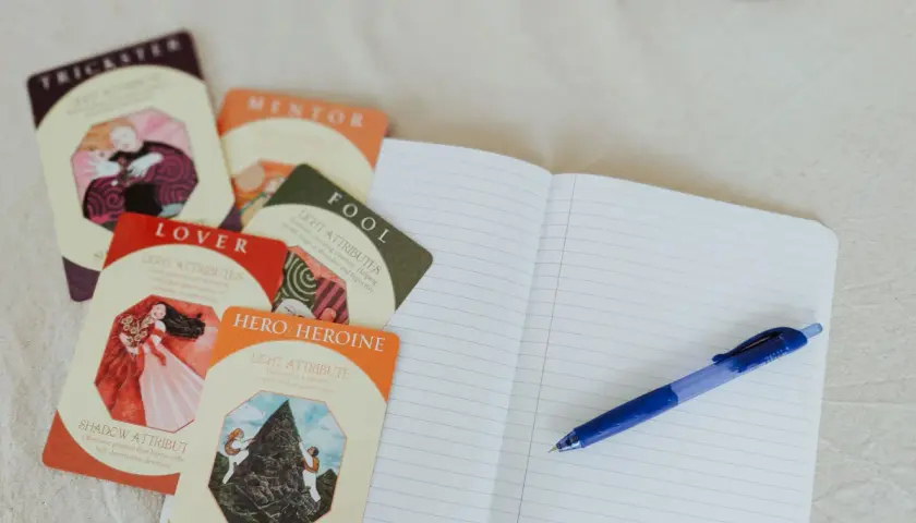 Cartes d'archétypes (Trickster, Lover, Hero/Heroine) et un carnet ouvert avec un stylo, pour une idée de nom pour personnage de roman.