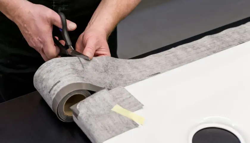 Hände schneiden Dichtungsband zu, um eine Duschwanne abzudichten. Das Material ist auf einer Rolle und wird präzise zugeschnitten.