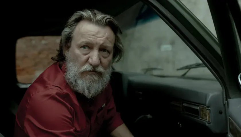 Wzgórze psów obsada: Ian McShane w czerwonej koszuli siedzi w samochodzie, z siwą brodą i zmęczonym spojrzeniem.