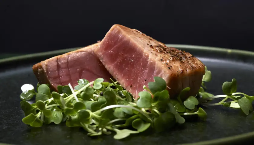 Två bitar perfekt grillad tonfisk, kryddad med peppar, vilar på en bädd av gröna skott på en mörk tallrik.
