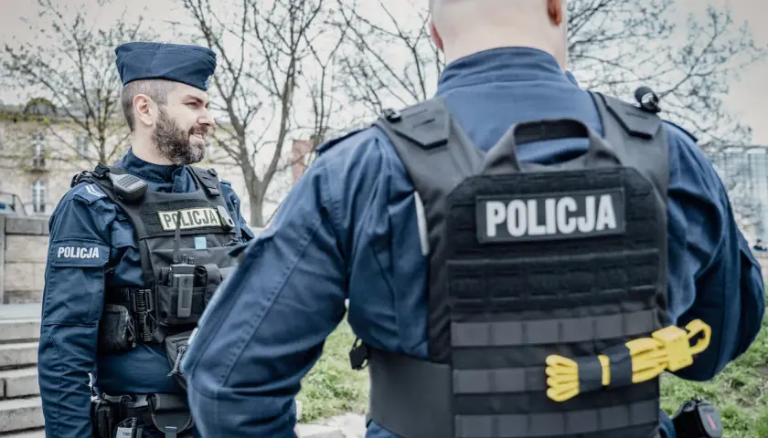 Dwóch policjantów w mundurach i kamizelkach taktycznych. Widoczne wyposażenie policjanta: radiostacja, kabura na broń, pas z ładownicami.