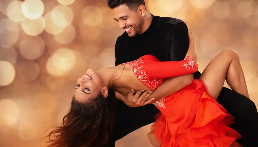 Coppia che balla con passione una salsa ballo latino, lei in abito rosso, lui in nero, su sfondo sfocato con luci dorate.