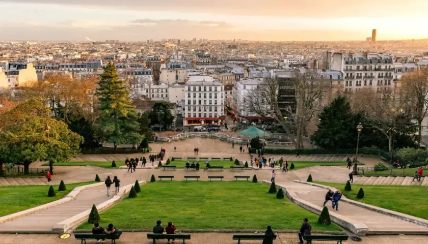 Widok z wzgórza Montmartre na Paryż. Ludzie spacerują po zielonych tarasach i odpoczywają na ławkach, podziwiając panoramę miasta.