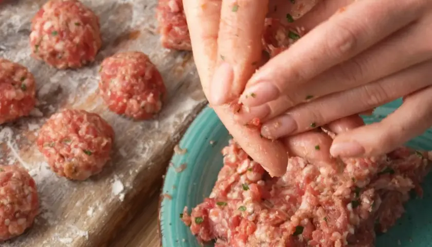 Ręce formują klopsiki według przepisu na meatballs. Na desce widać już gotowe, obtoczone w mące kulki mięsa.