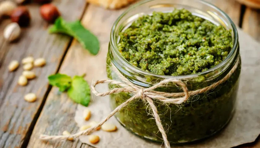 Słoik domowego pesto z bazylii, ozdobiony sznurkiem. Obok orzeszki piniowe i listki bazylii.
