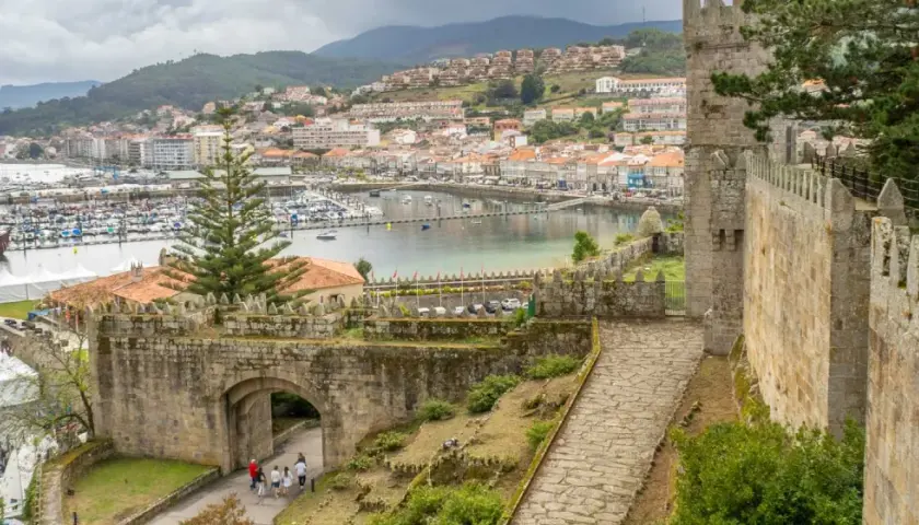 View from a castle wall overlooking a harbor filled with boats, a historic ship, and a town. The path leads through an ancient stone archway, hinting at a journey from porto to Baiona.