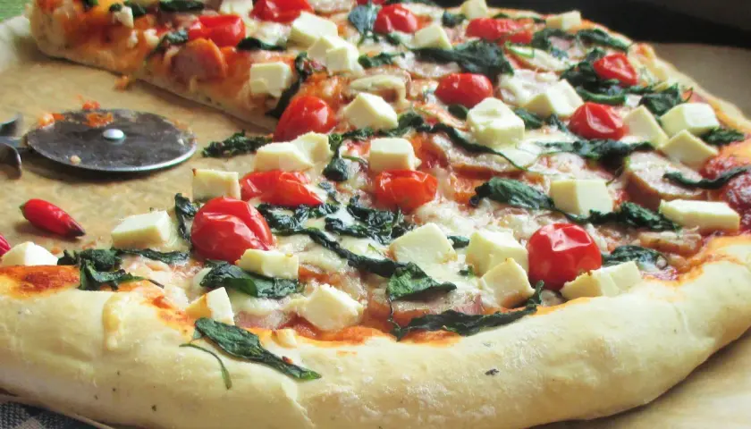 Pyszna pizza ze szpinakiem, pomidorkami koktajlowymi i kostkami sera feta. Obok leży nóż do pizzy i mała papryczka chili.