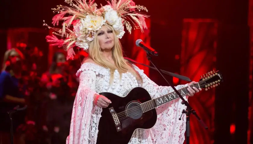 Artystka w białej, koronkowej sukni i kwiecistym wianku gra na gitarze. Jej muzyka sprawia, że pokonamy fale wykonawcy.