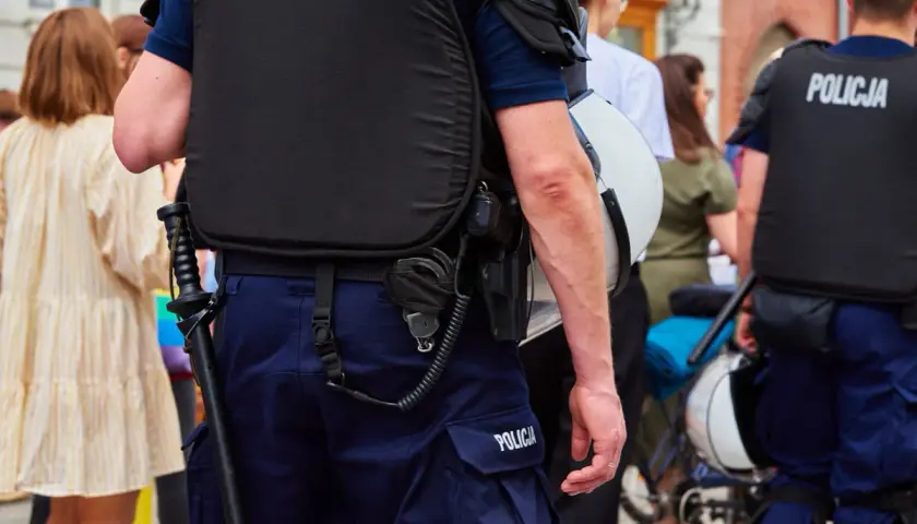 Funkcjonariusz publiczny w kamizelce taktycznej i mundurze, z pałką i kajdankami, stoi na straży porządku.