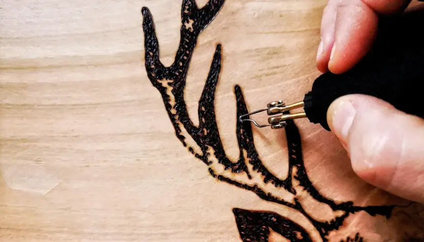 Dettaglio di lavori con pirografo su legno: una mano che disegna un palco di cervo con precisione.