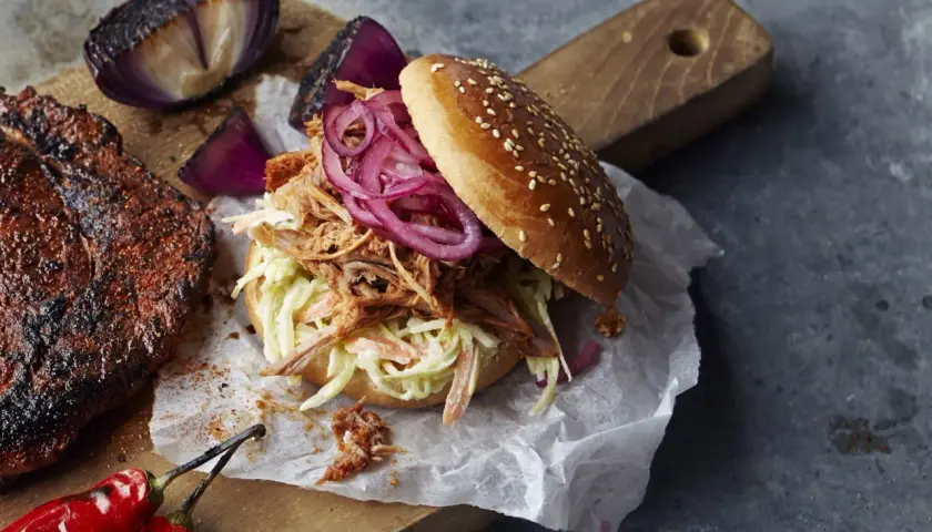 En saftig pulled pork-burgare med krispig coleslaw och rödlök, perfekt för ett bbq-recept. Bredvid ligger en bit grillat kött och rostade lökar.