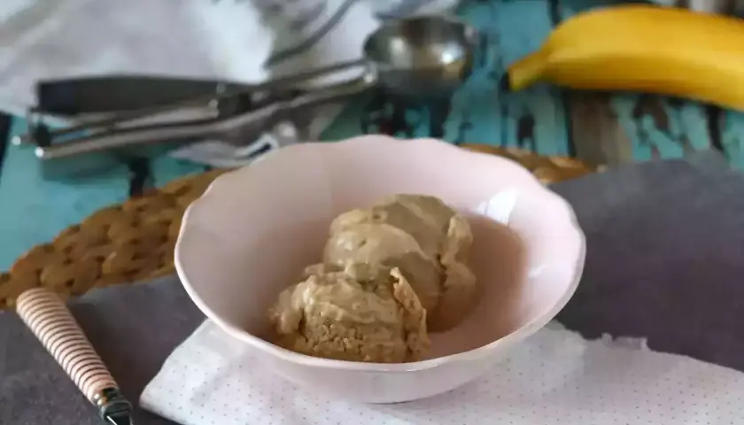 Zwei Kugeln cremiges, veganes Bananeneis in einer rosa Schale, daneben ein Löffel und ein Stück Gebäck. Eine Banane liegt im Hintergrund.