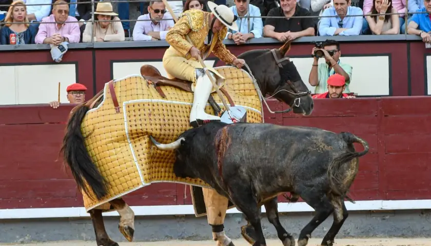 Un torero a caballo, con traje de luces, se enfrenta a un toro en la arena. El caballo lleva una manta amarilla. El público observa desde las gradas.