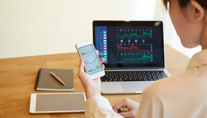 Frau analysiert Aktienkurse auf Laptop und Smartphone. Sie plant, wie sie ihre Aktiengewinne versteuern kann, während sie Notizen macht.