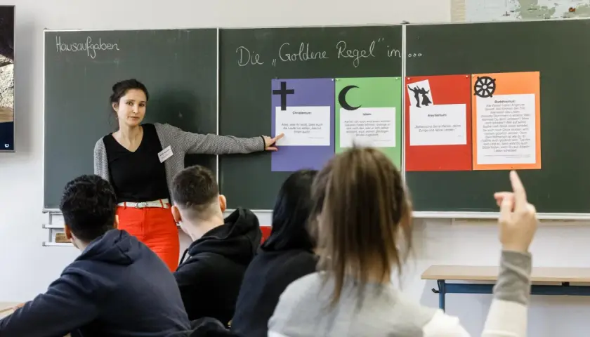 Lehrerin erklärt die Goldene Regel im Religionsunterricht Grundschule. Tafeln zeigen Zitate aus Christentum, Islam, Alevitentum und Buddhismus.