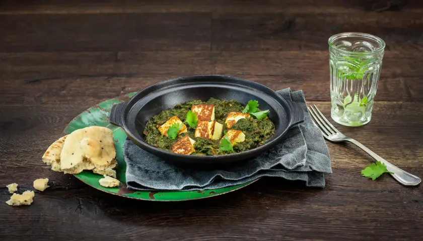 En skål med krämig palak paneer, toppad med gyllene paneerbitar och färsk koriander. Serveras med naanbröd och en glasdryck.