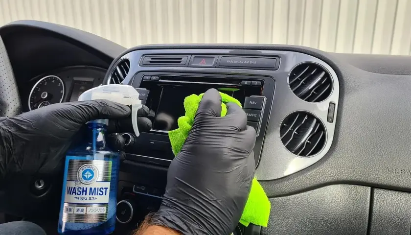 Ręce w czarnych rękawiczkach czyszczą deskę rozdzielczą auta zieloną ściereczką i preparatem Wash Mist. Prosty sposób, jak odnowić plastiki w aucie.