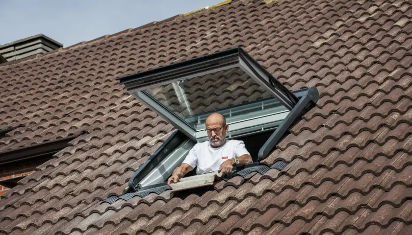 Mężczyzna montuje okno dachowe balkonowe na brązowym, dachówkowym dachu.