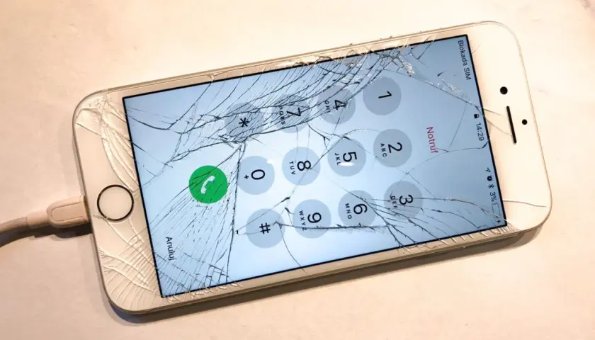 Zepsuty ekran telefonu z widocznym pęknięciem, podłączony do ładowarki. Na ekranie widoczna klawiatura numeryczna.