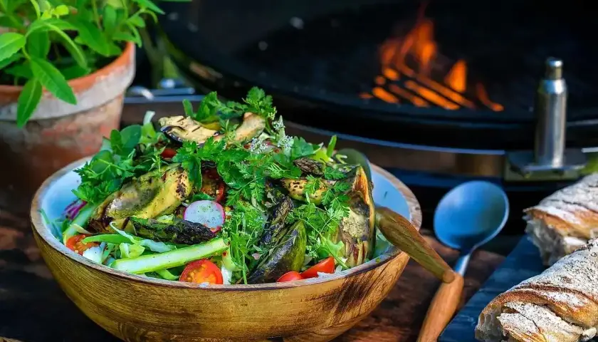 En stor skål med god sallad, grillad aubergine och sparris, med en grill i bakgrunden. Perfekt för en sommarkväll.