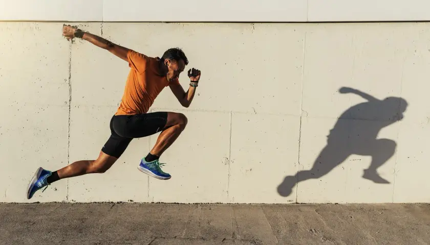Biegacz w pomarańczowej koszulce i czarnych spodenkach w dynamicznym skoku, jego cień odbija się na ścianie. Trening na świeżym powietrzu, by poprawić VO2 max.