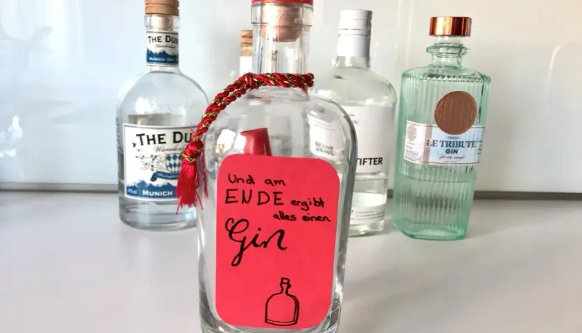 Ein roter Zettel mit der Aufschrift "Und am Ende ergibt alles einen Gin" ist an einer leeren Flasche befestigt. Mehrere Gin-Flaschen stehen im Hintergrund.