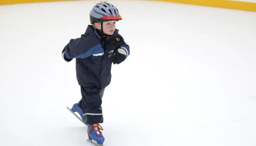 Bambino con casco e tuta blu pratica il pattinaggio sul ghiaccio, con i pattini azzurri e lacci arancioni.