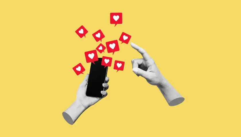 Dwie ręce, jedna trzyma smartfon, druga wskazuje na ekran, z którego wylatują czerwone dymki z serduszkami, symbolizujące polubienia w social mediach.