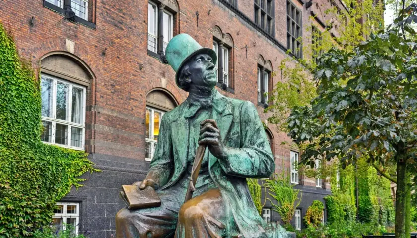 Pomnik Hansa Christiana Andersena w zielonej czapce, siedzącego z książką i laską, na tle ceglanego budynku.