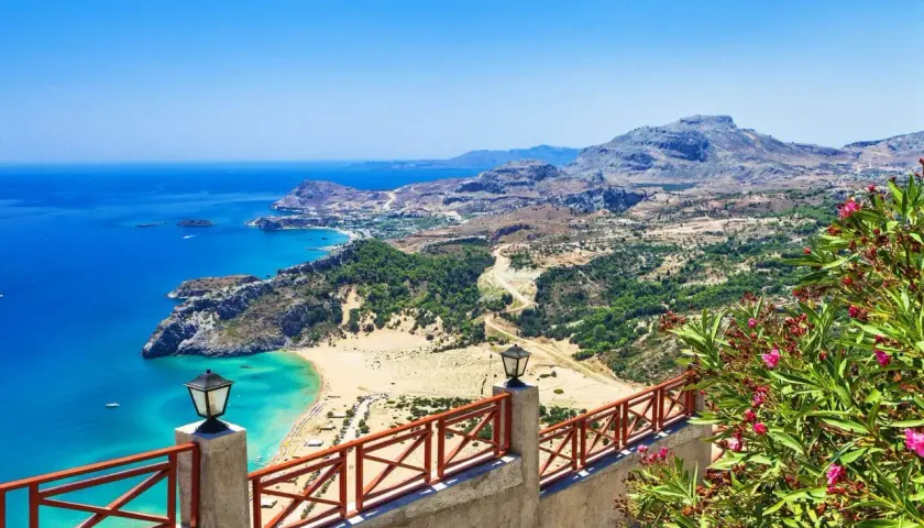Blick auf eine Bucht mit türkisfarbenem Wasser, Sandstrand und grünen Hügeln. Ein Geländer mit Laternen und blühenden Oleandern im Vordergrund. Rhodos Geheimtipps für unvergessliche Ausblicke.